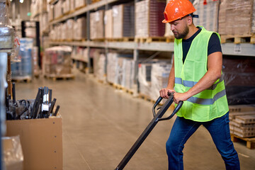male staff worker in warehouse uses hand pallet stacker to transport goods, alone, dressed in working clothes and safety hard hat.Skilled warehouse employee pushing manual pallet jack