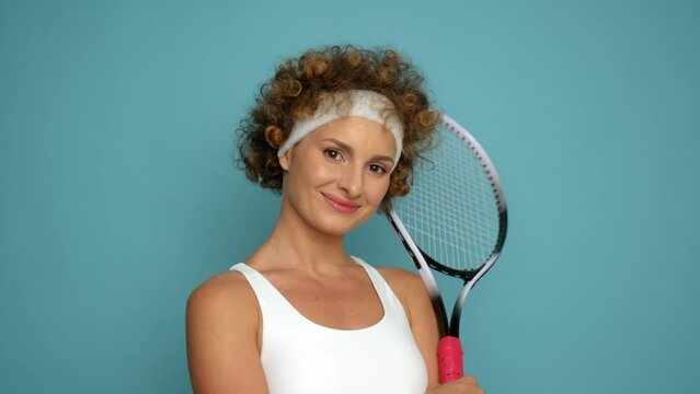 Curly-haired beauty stands in nice pose on blue background in the studio, smiling and pressing tennis racket to herself with smile, sport style concept, copy space
