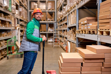 pleasant warehouse employee pushing manual pallet jack and working in factory storage room, warehouse worker transporting cardboard goods delivery. logistics and distribution concept