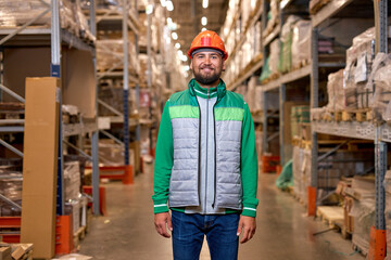 Portrait of happy smiling young warehouse worker posing posing near shelves with goods. Feeling satisfied doing job good. Young employee in working uniform and safety helmet, look at camera