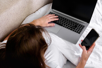 A young woman is working at a laptop and with mobile phone on a white bed.