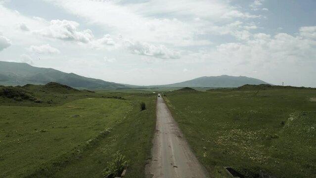 Drone video. Flying over an old road behind the car between green hills covered with grass. A village appears from behind a hill against a background of high mountains in sunny weather. Armenia.