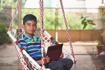 Indian little boy using Tablet resting in swing hammock outdoors	
