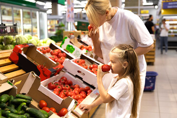 Shopping with kids. Mother and child buying fresh vegetables on a market. Mom and little girl daughter smell tomatoes in the store before buying. Family in shop. Parent and children in a mall - obrazy, fototapety, plakaty