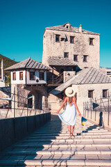 Woman tourist in white dress and hat crossing old bridge in Mostar- Bosnia