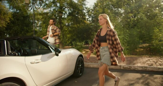 A guy and a girl in plaid shirts walk down the street and get into a white convertible in a park in summer. A brunette guy and a blonde girl get into a white car and leave the park
