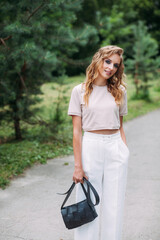 A young slender girl with fashionable make-up and a stylish hairstyle, in white trousers and a beige top, with a black leather handbag in her hands, stands against the background of the park.