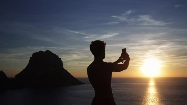 Shirtless man enjoying the view and taking a panoramic photo of the sunrise.