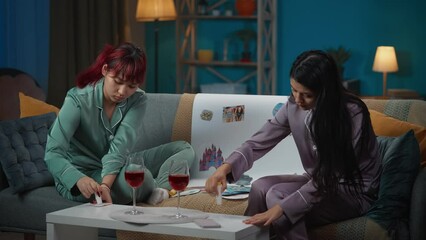 Medium-full video capturing two young women wearing pajamas sitting on the couch in a room, drinking wine and making a collage, moodboard with their wishes.