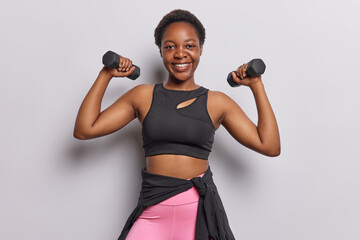 Cheerful young dark skinned woman lifts dumbbells has strength workout does biceps exercises...