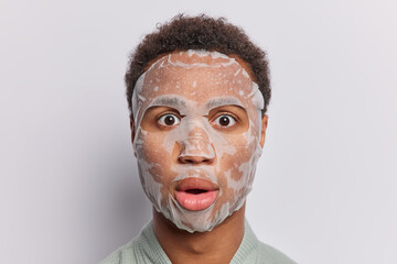 Headshot of shocked young man undergoes moisturizing procedure puts on beauty sheet mask on face...