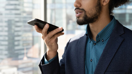 Close up cropped of African American businessman executive in suit recording audio voice message on smartphone, holding phone near mouth, dictating, chatting online by speakerphone in social network