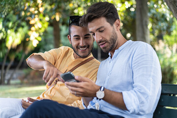Two Men Discussing Smartphone Content in the Park - Two men in their 30s, wearing shirts, sit on a park bench in summer shade. One holds a phone, the other, smiling, points at the screen.