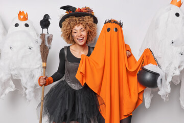 Halloween party. Cheerful young woman poses with orange ghost and broom prepares for special holiday wears wizard hat and black dress isolated over white background prepares for house party.