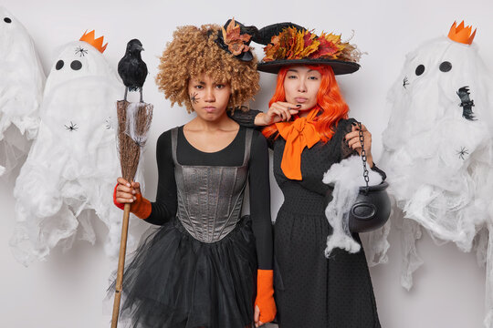 Two serious women dressed in black clothes pose with halloween attributes express sorrow focused at camera prepare for carnival on 31st of October surrounded by spooky ghosts. Magical festivities