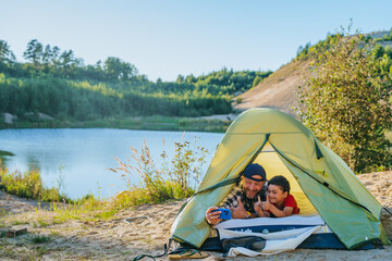 Smiley bearded European man laying in tent with little son using phone, makes video call. Cute  preschooler boy Showa thumb up gesture for grandparents via internet chat. Outdoor leisure, parenthood. © Iona