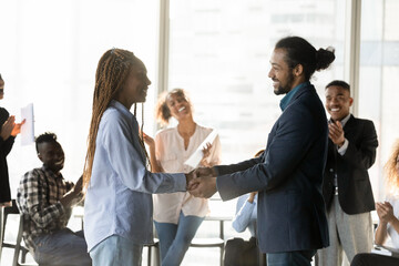 Smiling African American business partners shaking hands at meeting side view, colleagues applauding on background, executive team leader congratulating employee with promotion at briefing