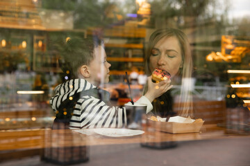 Shot through window. Happy blonde mom and her little funny son rest in the cafe, drink tea and eat...