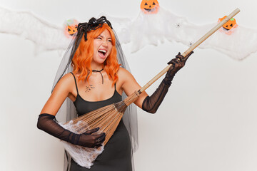 Portrait of a happy young woman in halloween costume holding broom as a guitar and playing on the rock concert, smiling isolated over white background
