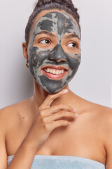 Vertical shot of cheerful dark haired woman touches chin gently applies nourishing clay mask for skin treatment focused aside wrapped in bath towel isolated over white background. Beauty procedures