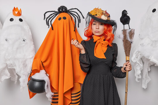 Magic and spell. Studio photo of young concentrated European female wearing costume of witch celebrating Halloween organising party posing among scary white and orange ghosts with red long hair