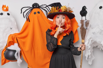 People and holiday celebration concept. Indoor shot of young European female in Halloween party wearing costume of witch standing surrounded by spooky ghosts making gesture to be silent and keep still