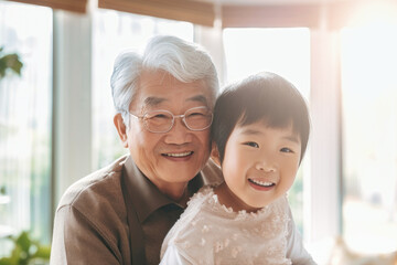 An elderly man with a little Asian girl in the room. They hug, have fun and rejoice at the meeting. Meeting of granddaughter and her grandfather. Caring for the elderly. Family values. - obrazy, fototapety, plakaty