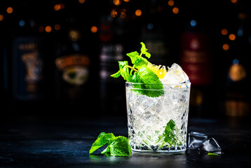 Tommy's margarita summer cocktail drink with tequila, lime juice, lemon, syrup, mint and ice in glass, dark background, bar tools and bottles