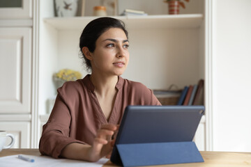 Dreamy Indian woman distracted from tablet, looking in distance, sitting at desk with modern electronic device, pensive young businesswoman or student visualizing good future, dreaming of success