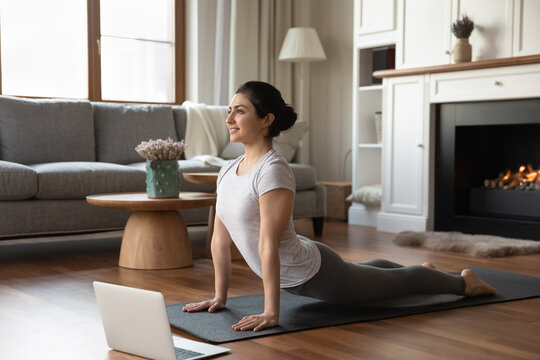 Smiling Indian Woman Practicing Yoga Online Using Laptop, Working Out At Home, Happy Young Female In Grey Sportswear Doing Upward Facing Dog Pose, Stretching In Urdhva Mukha Shvanasana Exercise