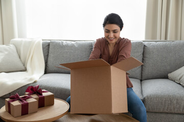 Smiling Indian woman unpacking parcel with wrapped gift boxes, sitting on couch at home, smiling young female opening unboxing birthday presents, package with surprises, excited by delivery - obrazy, fototapety, plakaty