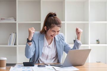 Asian businesswomen show joyful expression of success at work smiling happily with a laptop computer in a modern office.