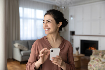 Dreamy smiling Indian woman holding smartphone, looking in distance, standing at home, happy pensive young female distracted from online chatting, visualizing good future, planning and dreaming