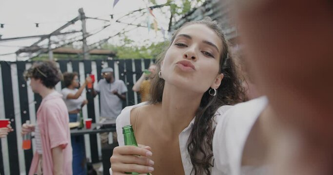 Slow motion portrait of joyful young woman posing for selfie with beer bottle at summer party outside. Lady looking at camera smiling holding alcohol while friends dancing in background.