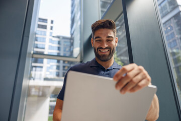 Fototapeta premium Handsome smiling freelancer working on laptop while standing in coworking near windows