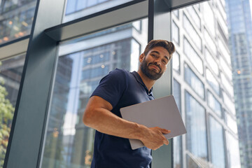 Fototapeta premium Handsome freelancer with laptop standing in coworking near window and looking at camera