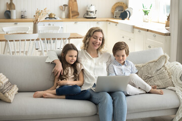 Smiling mother and cute kids sitting on cozy couch with laptop, look at device screen, smile, spend weekend time at home, having fun watching cartoons or family movie, browse internet use application