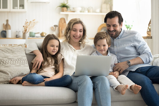 Happy attractive parents and little children sitting on couch looking at computer screen, watch funny cartoons, smile, have fun together using modern technology, chatting online, buying on internet