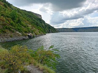 A view from the thicket of coastal vegetation to the wide water surface of the Dniester river bed, covered with a small ripple that reflects sunlit clouds.