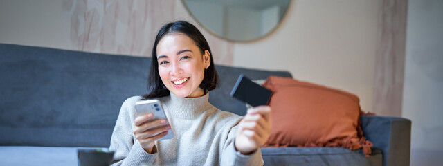 Smiling asian girl with smartphone and credit card, does her shopping online, uses mobile phone to...