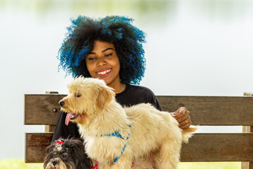 Jovem mulher negra com cabelos pintados de azul, com seus dois cachorros em um banco do parque.