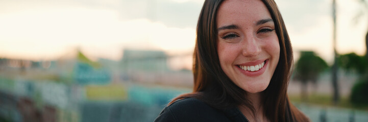 Portrait of cute girl with freckles and loose brown hair wearing black sports hoodie looks at the camera and walks on the bridge of the modern city background