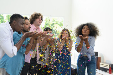Group of six friends having fun at home blowing colorful confetti. Happy young people celebrating...