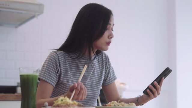 joyful nice Asian Woman having asian noodles food pad thai holding chopsticks for lunch during lunch break and typing in her smartphone or browsing social networks on the table in kitchen at home