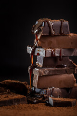 Fototapeta premium A pile of dark broken chocolate sprinkled with cocoa powder and poured with chocolate sauce on a dark background