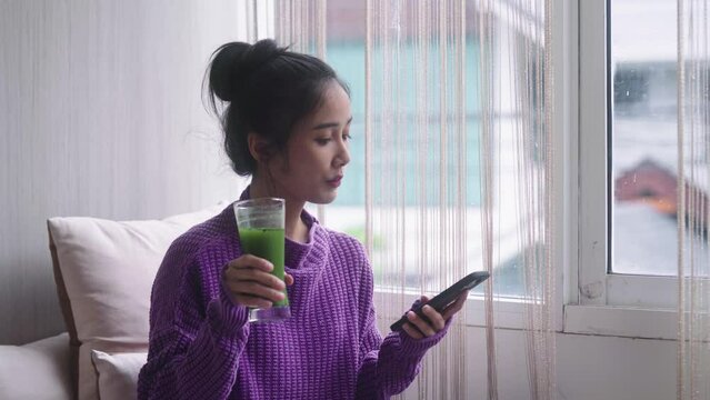 Young happy millennial healthy Asian woman checking smartphone while drinking homemade vegan detox juice at home, healthy dieting food concept.
