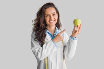 Cheerful young lady doctor in uniform pointing finger on apple isolated on light grey background,...