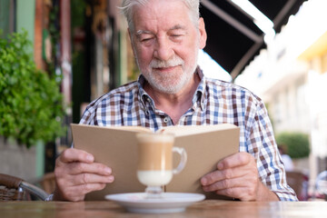 Portrait of bearded senior man sitting outdoor at a cafe table reading a book while enjoying a coffee and milk drink - caucasian elderly man relaxed in retirement or vacation