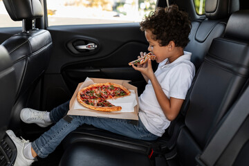 Hungry black boy sitting on car back seat, eating pizza