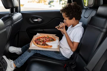 Poster Hungry black boy sitting on car back seat, eating pizza © Prostock-studio